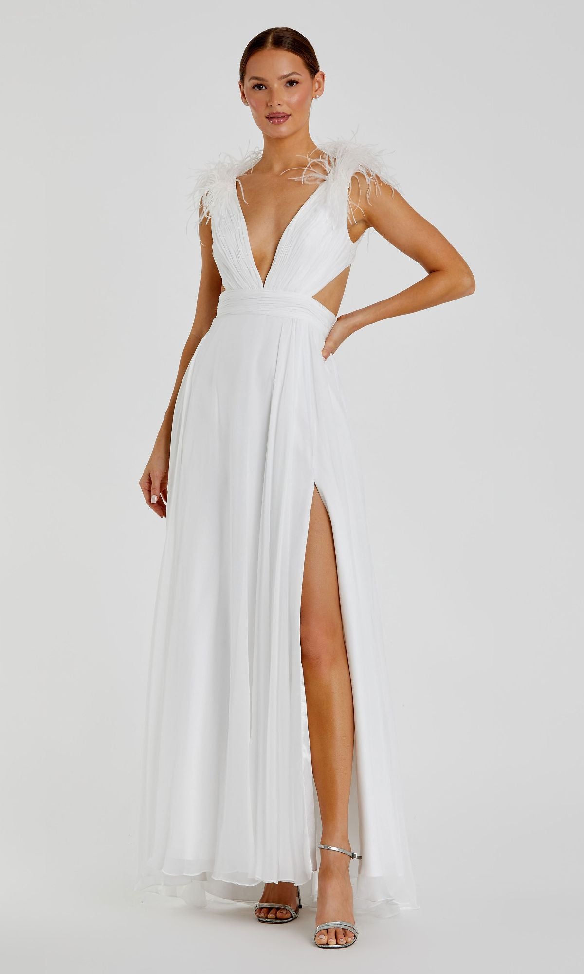 A woman models the Mac Duggal 68113 Feather-Trim Long Formal Dress—a sleeveless white gown with feathered shoulders, deep V-neck, high slit—paired with silver high heels, standing against a plain background.